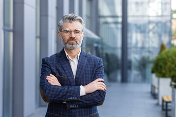 Portrait of mature successful boss, gray haired businessman looking at camera with crossed arms, man outside office building in business suit and glasses, satisfied and focused banker investor.