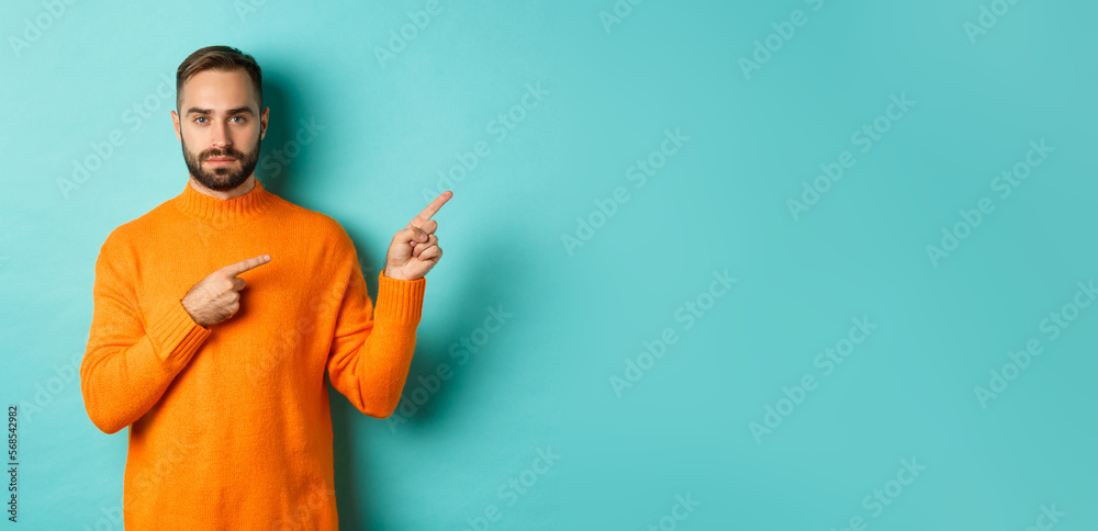 Wall mural Handsome and serious bearded man in orange sweater pointing right, showing advertisement or logo, standing over turquoise background