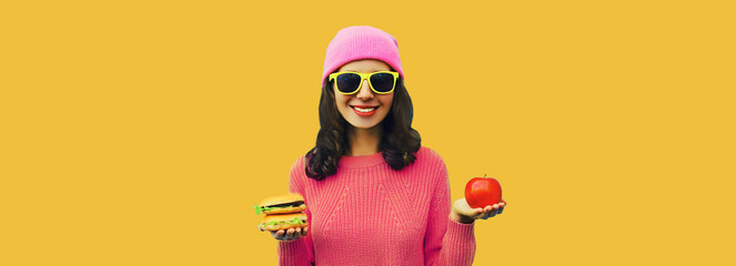 Portrait of stylish happy smiling young woman making choice showing burger fast food and apple on yellow colorful background