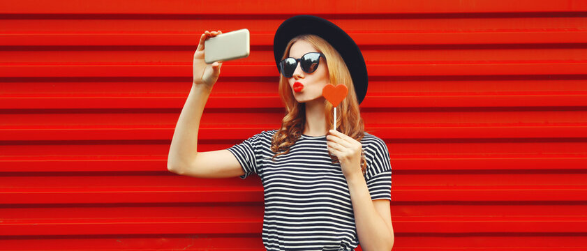 Portrait of beautiful young woman taking selfie with smartphone blowing her lips sends air kiss holding red heart shaped lollipop wearing black round hat on background