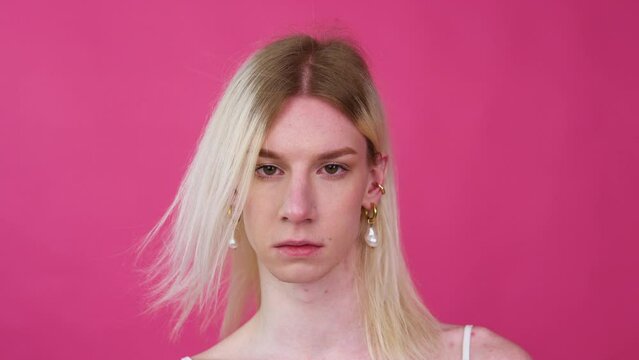 Portrait of young transvestite man in a dress puts on earrings on a pink background