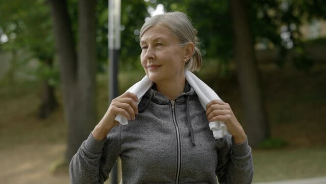 Portrait of the charming mature woman in sporty clothes carrying towel in her neck. Elderly female looking at camera before the training standing in park.