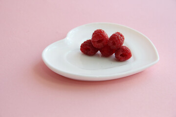 ripe raspberries on a white plate in the shape of a heart on a pink background. raspberry berry on a plate