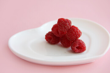 ripe raspberries on a white plate in the shape of a heart on a pink background. raspberry berry on a plate