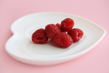 ripe raspberries on a white plate in the shape of a heart on a pink background. raspberry berry on a plate