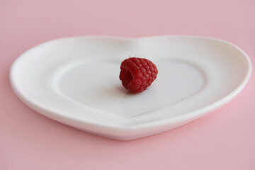 ripe raspberries on a white plate in the shape of a heart on a pink background. raspberry berry on a plate