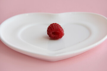 ripe raspberries on a white plate in the shape of a heart on a pink background. raspberry berry on a plate