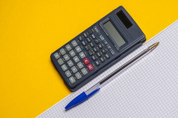 Calculator with squared notebook and ballpoint pen (biro) for math, physics, engineering or business homework and tax and finance calculations on a yellow background. Copy space.
