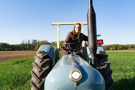 Junge Frau mit Spass an der Landwirtschaft sitzt auf einem Oldtimer - Traktor mitten im Getreidefeld bei Sonnenschein.
