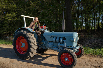 Junge Frau mit Spass an der Landwirtschaft sitzt auf einem Oldtimer - Traktor auf einem Feldweg. 