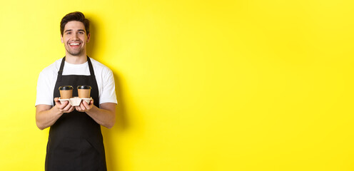 Friendly barista in black apron giving takeaway order, holding two cups of coffee and smiling, standing over yellow background
