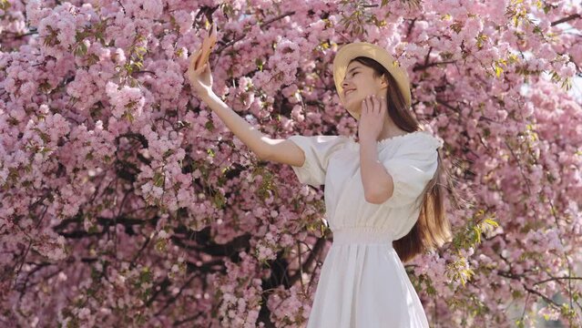 Attractive young woman in straw hat and white summer dress using modern smartphone for taking selfie near sakura tree with pink flowering. People and technology concept