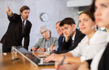 Dissatisfied male boss telling off office workers of different ages using notebooks at table in office