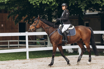 A girl rides fast on a brown racehorse next to a hurdle at a show jumping competition on a clear...