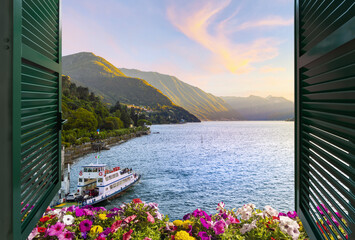 View from a balcony terrace with flowers and shutters of the mountains and lakefront promenade on Lake Como at the town of Bellagio, Italy, under a colorful sunset. - Powered by Adobe
