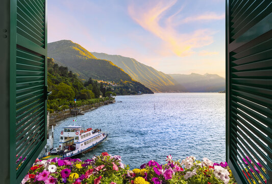 View from a balcony terrace with flowers and shutters of the mountains and lakefront promenade on Lake Como at the town of Bellagio, Italy, under a colorful sunset.