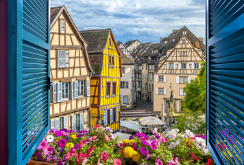 View from an open window overlooking a small square with cafes and half timber frame buildings in the medieval historic center of Colmar, France.