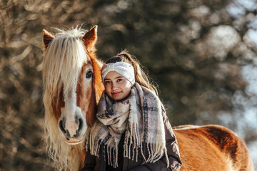 Cute equestrian and horse friendship scene: A young teenage girl interacting with her haflinger horse in front of a snowy rural landscape in winter outdoors