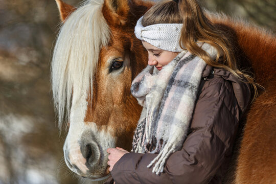 Cute equestrian and horse friendship scene: A young teenage girl interacting with her haflinger horse in front of a snowy rural landscape in winter outdoors