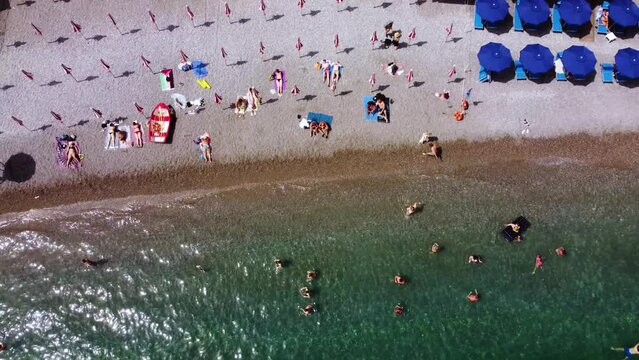 Aerial drone footage of the Marina Grande beach, town hall, Sant'Andrea duomo bell tower in Amalfi coast, Campania, Italy. Parasol, sun loungers, people swimming and colourful cliff villas from above.