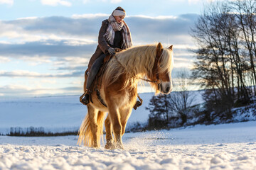 A young equestrian teenage girl rides on her haflinger horse through the snow in the evening during sundown in front of a snowy rural winter landscape