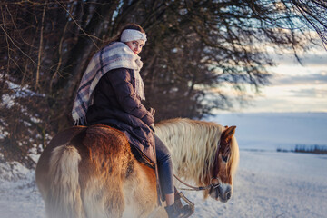 A young equestrian teenage girl rides on her haflinger horse through the snow in the evening during...