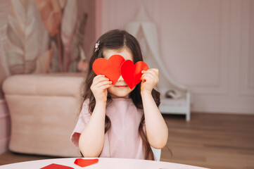 a little girl makes Valentine's Day cards using colored paper, scissors and pencil, sitting at a table in a cozy room.