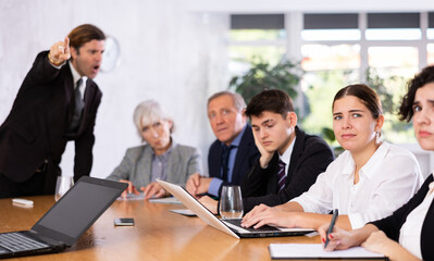 Obraz premium Upset young girl sitting with colleagues at table in office meeting room scolded by angry boss expressing dissatisfaction with work of subordinates..