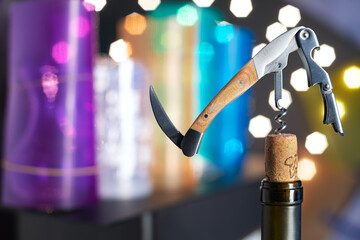 folding corkscrew and the neck of a wine bottle with a cork against the background of multi-colored lights. Blurred background. Space for text.