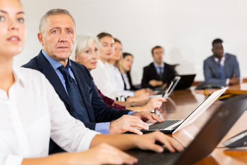 Serious late-middle-aged white businessman sitting among colleagues at conference table in meeting room and listening to presentation