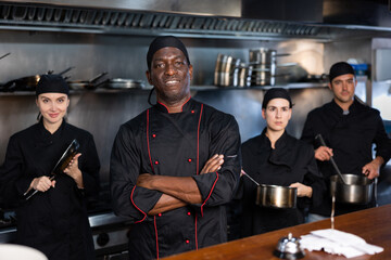 Fototapeta premium Team of focused chefs with African man head chef posing together in restaurant kitchen