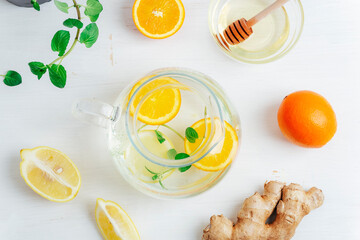 Glass teapot with orange, lemon, ginger and mint tea with ingredients on white wooden table. Top...