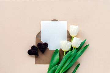 White tulips, envelope with blank sheet and chocolate hearts on neutral beige background. Valentine's Day, Mother's Day holiday concept. Top view, flat lay, mockup
