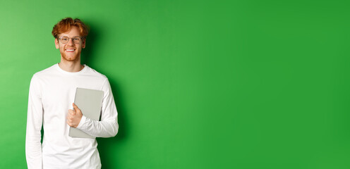 Handsome young man with red hair, wearing glasses and long-sleeve t-shirt, holding laptop on green background