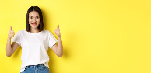 Pretty young asian woman in white t-shirt, showing thumbs up and smiling, praise good offer,...