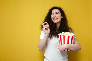 young cute girl viewer in a white t-shirt holds a big bucket of popcorn, smiles on a yellow background