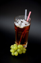 A tall faceted glass of fruit juice with ice and straws on a black background, next to a bunch of ripe green grapes.