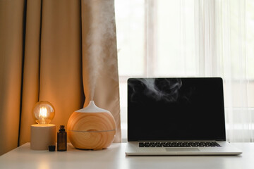 Aroma oil diffuser in work place, laptop and home decor on the table against window.