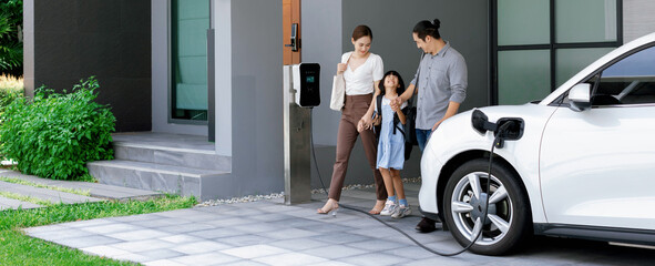 Fototapeta Progressive young parents and daughter with electric vehicle and home charging station. Green and clean energy from electric vehicles for healthy environment. Eco power from renewable source at home. obraz