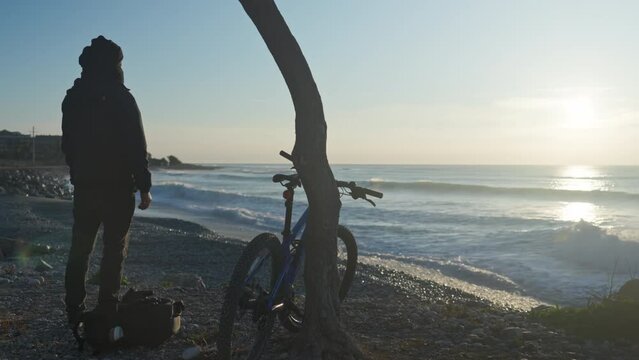 A solitary man stands on a gravel beach, watching the sunrise over the sea. His bag is laid beside him and his bike leaned against a nearby tree. This is a perfect moment of reflection and tranquility