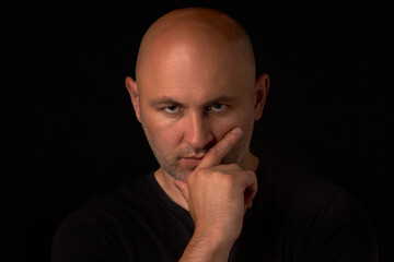 Bald, bearded man looks thoughtfully into the camera and holds his hand on his chin, on a black background. A man in depression, a midlife crisis