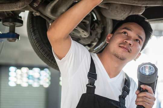 Portrait Of Asian Mechanic Man Holding Flashlight Checking For Repair Under Car Maintenance. After Service At Auto Garage Shop