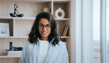 Attractive African American girl in casual looks at camera standing against book shelve home. Young Brazilian woman in glasses relaxing at hotel room. Successful fashion model indoors. Business.