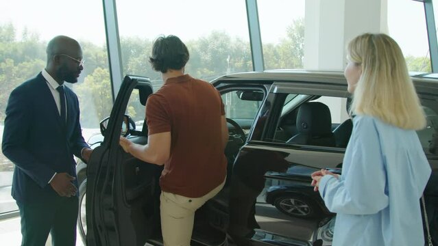 Black man wearing suit working in car dealership opening crossover car door and suggesting customers get in there