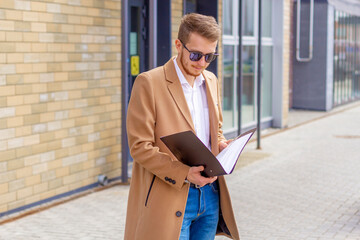 Fototapeta premium Portrait of a handsome man, businessman, scientist or teacher. He walks along a modern city street with a folder of documents and a notebook in his hands