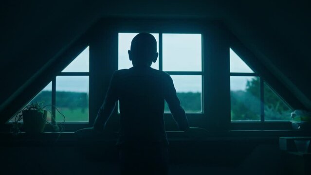 Silhouette of a young boy child looking out the window at dusk in a dark room before he goes to bed.