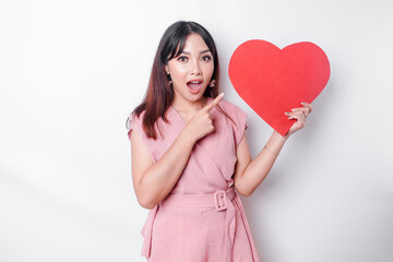 A shocked Asian woman dressed in pink, pointing at the copy space beside her while holding a big red heart-shaped paper, isolated by a white background. Valentine's Day concept.