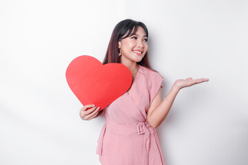 Excited Asian woman dressed in pink, pointing at the copy space beside her while holding a big red heart-shaped paper, isolated by white background