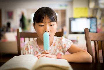 Young asian girl reading a book while licking popsicle