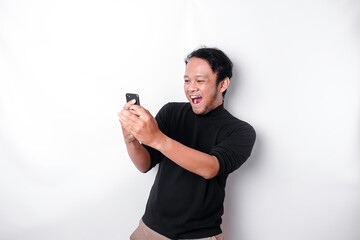 Fototapeta na wymiar A young Asian man with a happy successful expression wearing black shirt and holding his phone, isolated by white background
